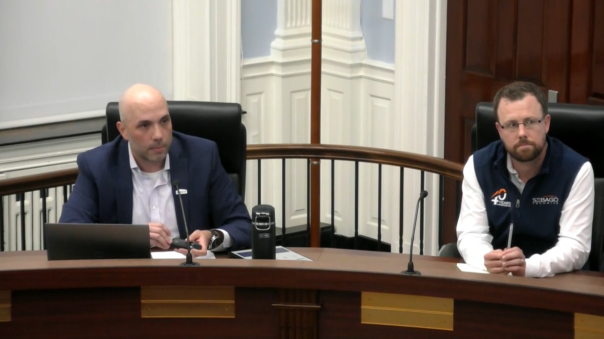 A bald man on the left in a suit and a bearded man on the right with glasses listen at a public meeting